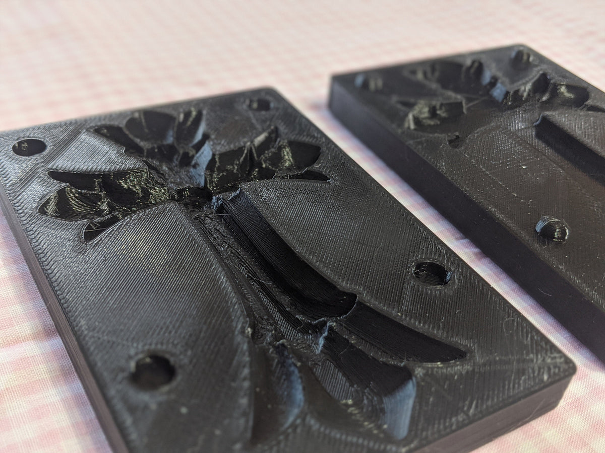 Two separated black Easter cross butter mold halves on pink gingham, with an ornate cross cavity and alignment holes.