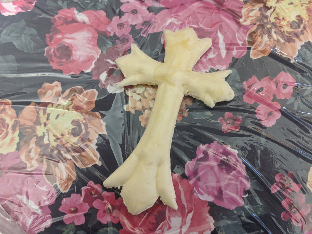 Overhead view of a plain pale yellow butter cross with flared upper arms and a split base on clear plastic over floral print.