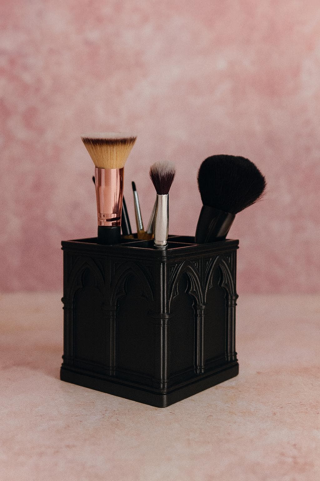 Black cathedral-arch makeup brush holder with four brushes against a pink background