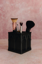 Black cathedral-arch makeup brush holder with four brushes against a pink background