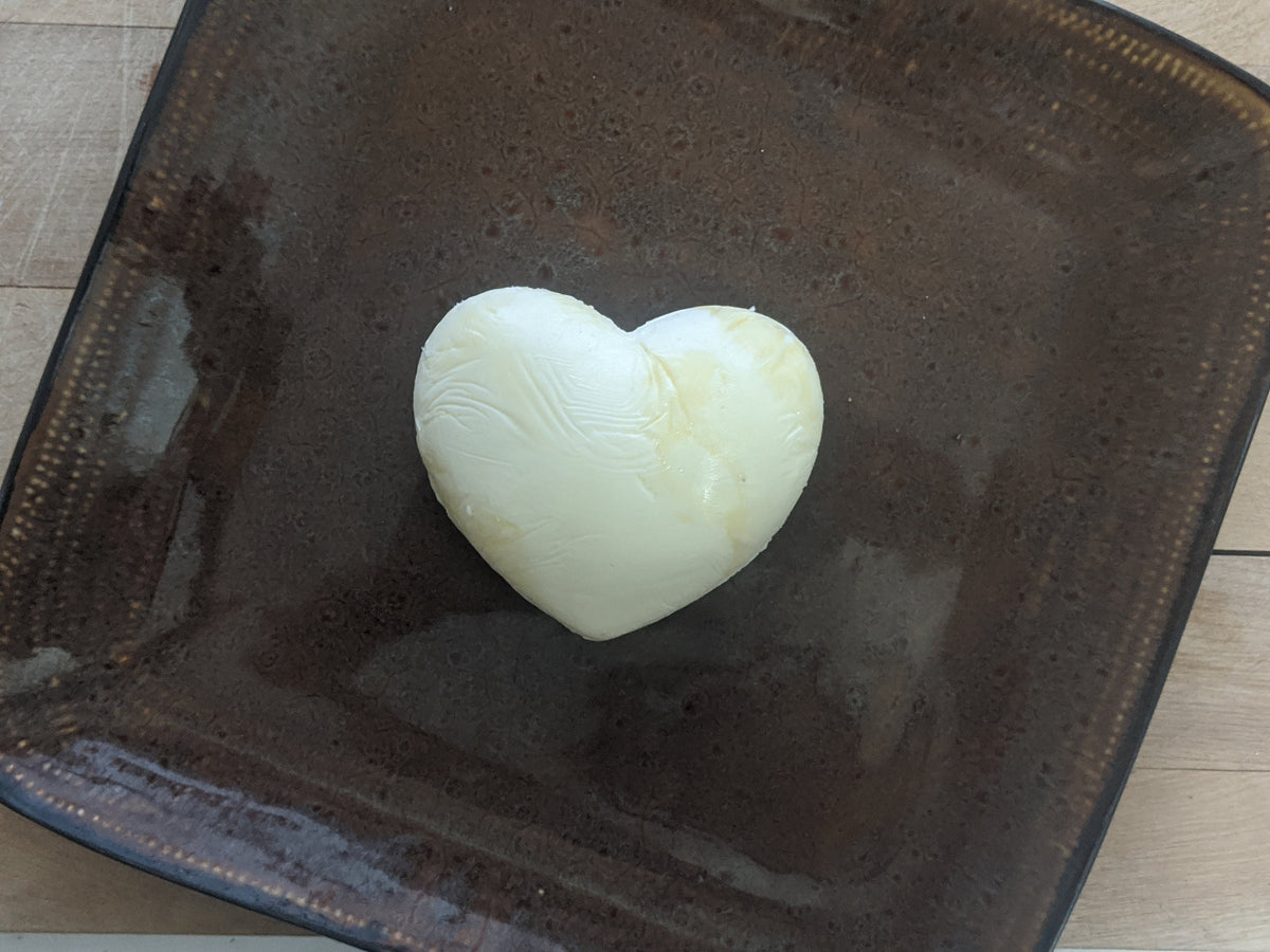 Plain pale heart-shaped butter pat centered on a dark brown square plate on a light wooden surface.