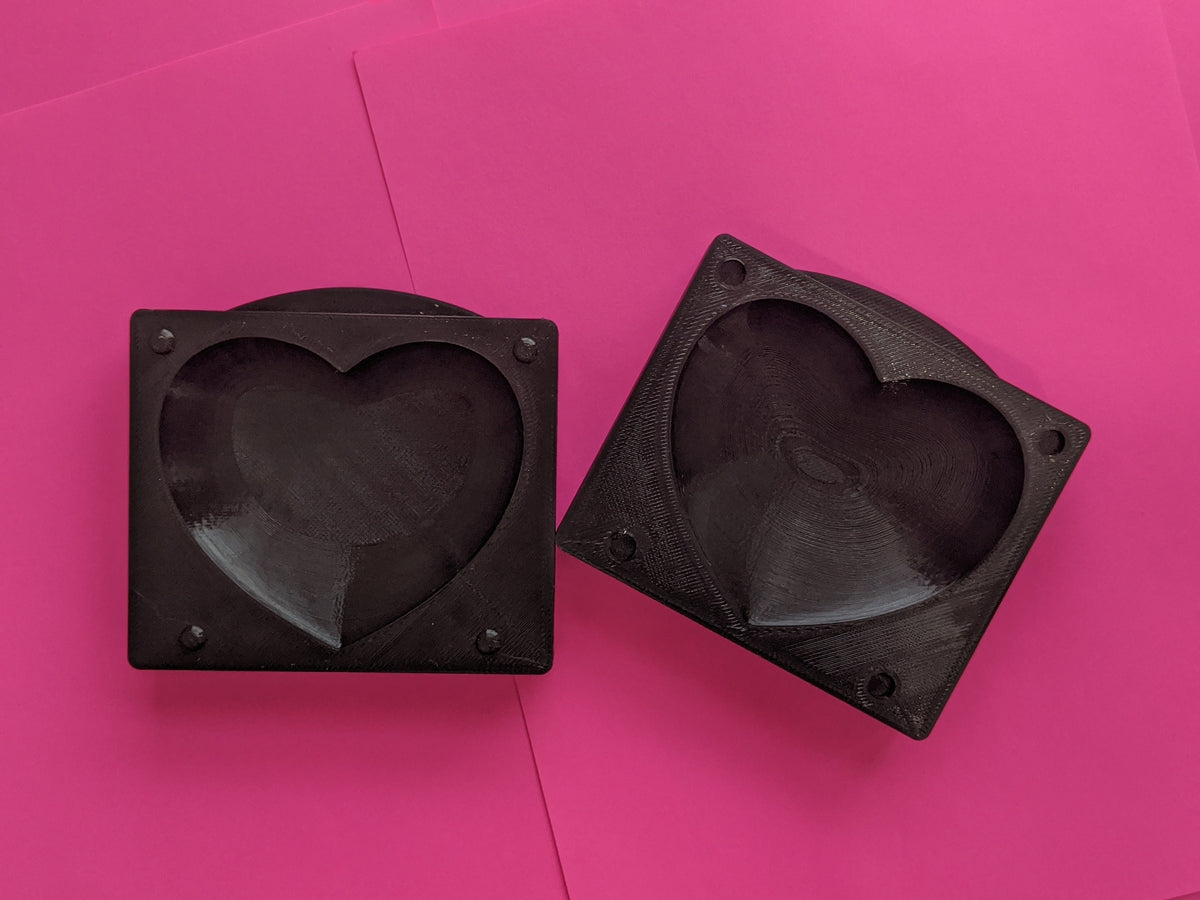 Two dark square heart butter mold halves on a bright pink surface, showing matching cavities and alignment features.