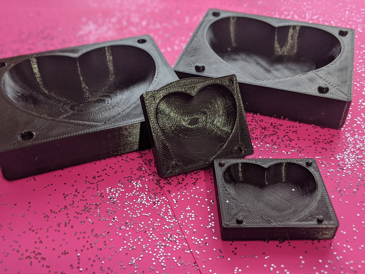 Dark heart mold components with rectangular blocks and square plates showing heart-shaped forms on a bright pink background.