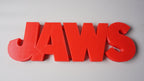 Close elevated front view of the red JAWS shelf sign showing rounded letter edges on a light-gray surface.