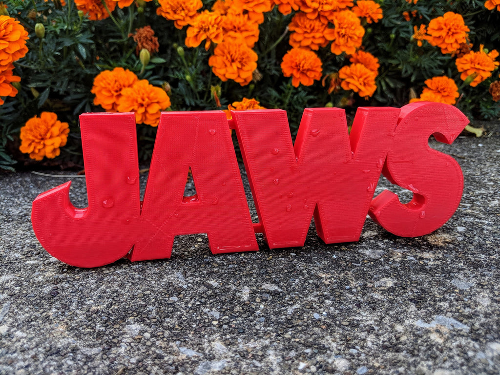 Red JAWS shelf sign standing outdoors on a rough speckled surface with green foliage and orange flowers.